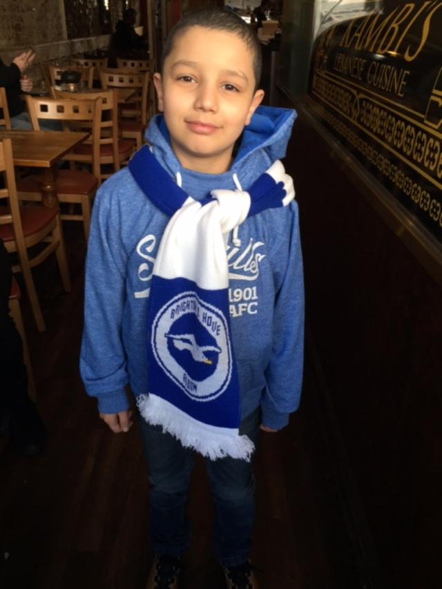 Daneal, a 8 year old boy, stands in a blue hoodie with a Brighton and Hove scarf