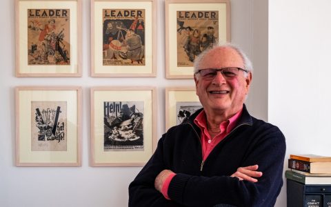 Paul, a white man with receding white hair, with an array of framed images from the Second World War, on the wall behind him