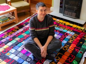 Dani, a white woman with short black hair, sitting on a colourful blanket in her living room
