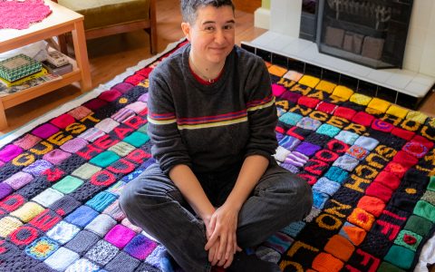 Dani, a white woman with short black hair, sitting on a colourful blanket in her living room