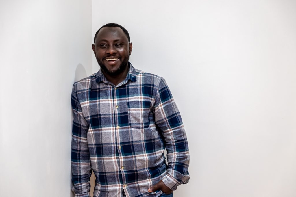 Luqman, a black man in his thirties, leaning on a white wall and smiling