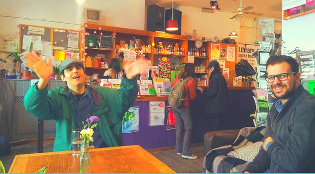 interior photo from the Jollof Cafe at the Cowley Club: two Middle-Eastern men sitting together at a table and and smiling, two other people in the background buying something at the counter