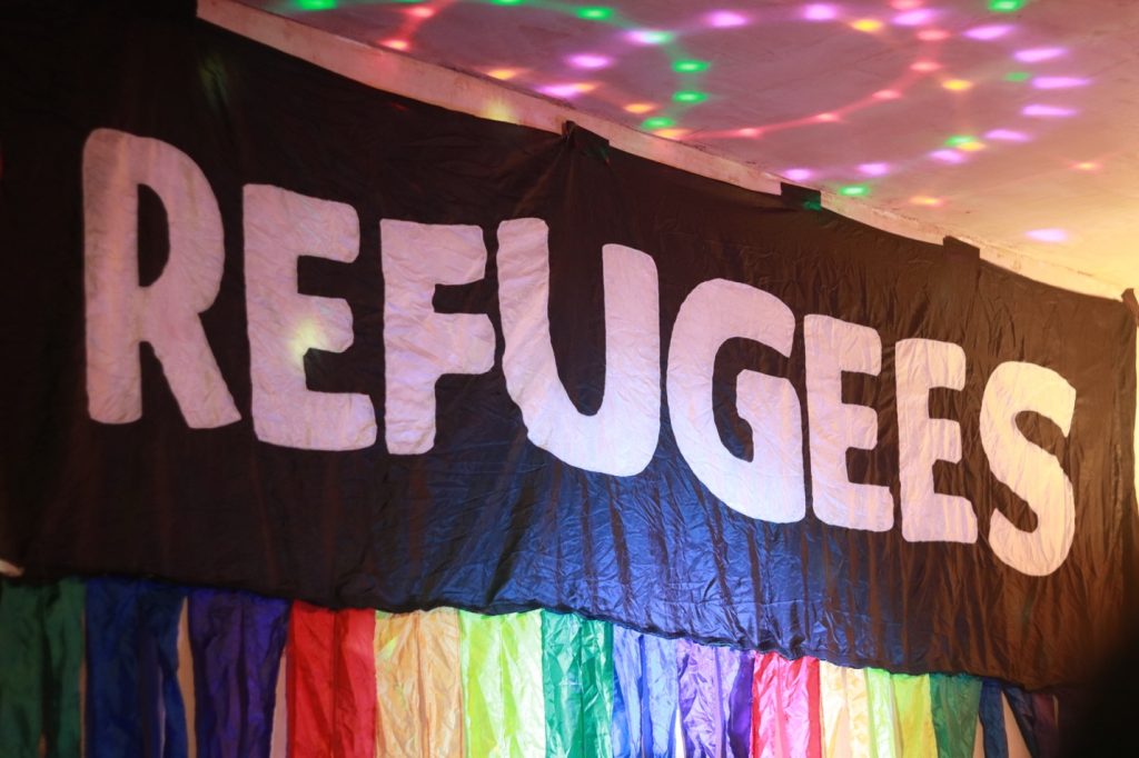 A black banner with large white letters spelling 'refugees'. The banner has a rainbow fringe