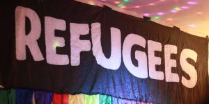 A black banner with large white letters spelling 'refugees'. The banner has a rainbow fringe