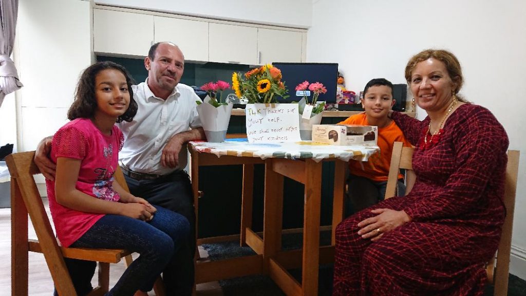 The family sit around their kitchen table in a semicircle. On the table there are flowers and a cake and a card which says "T4K thanks for your help. We shall never forget your kindness"