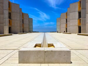 The Salk Institute, a concrete courtyard flanked by undulating concrete buildings. The lines draw the eye to the sea and sky beyond, Codera23, CC BY-SA 4.0 <https://creativecommons.org/licenses/by-sa/4.0>, via Wikimedia Commons” class=”wp-image-2951″/></figure></div>



<p style=