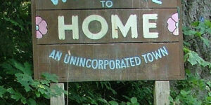 A wooden sign set against green ferns. Painted onto the weathered wood is the legend: Welcome to Home, an unincorporated town.