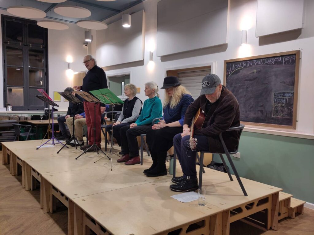 7 people in a row on a makeshift stage. The figure third from the left is standing, the others are seated. On the far right of the row a man holds a guitar