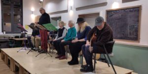 7 people in a row on a makeshift stage. The figure third from the left is standing, the others are seated. On the far right of the row a man holds a guitar