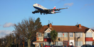 A BA jet flys low over a short parade of houses with cars parked outside. Trees and grass are visible