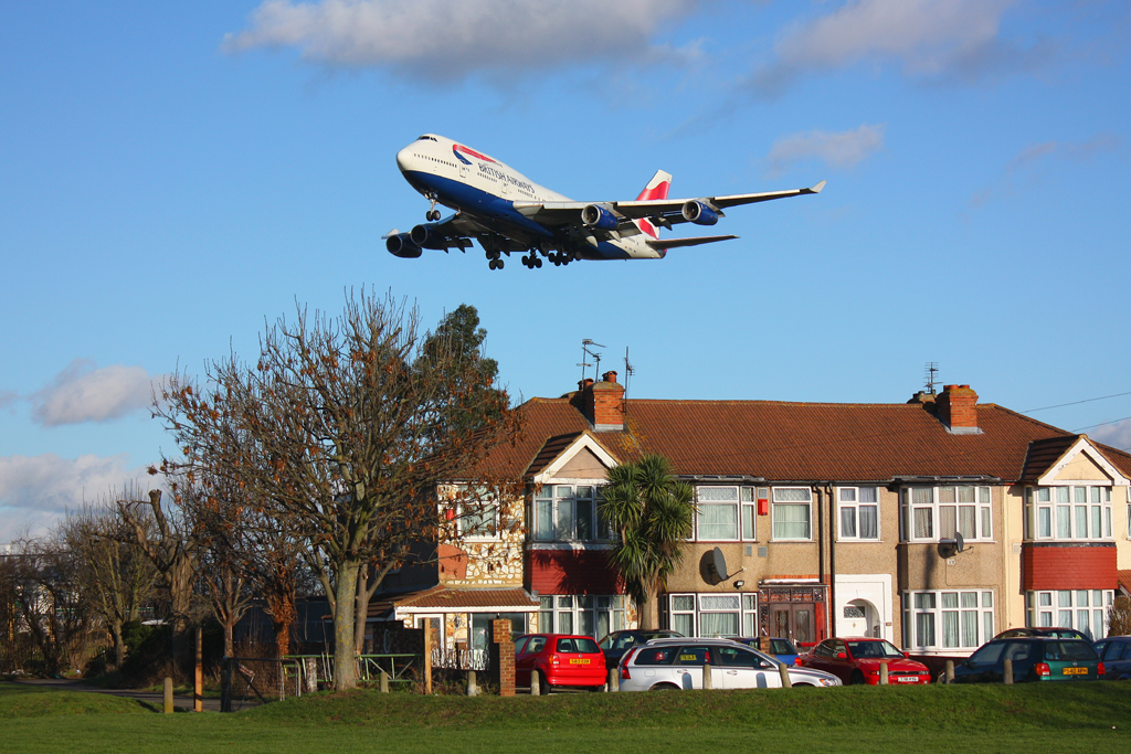 A BA jet flys low over a short parade of houses with cars parked outside. Trees and grass are visible