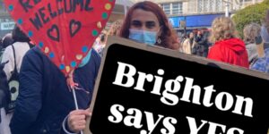 A woman in a blue jeans an a mask holds a red cardboard heart that says 'Refugees Welcome' and a sign that says 'Brighton says YES'. She is part of a small demonstration.