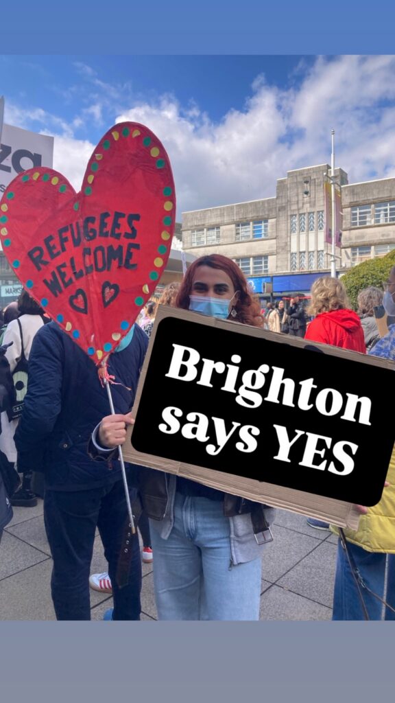 A woman in a blue jeans an a mask holds a red cardboard heart that says 'Refugees Welcome' and a sign that says 'Brighton says YES'. She is part of a small demonstration.